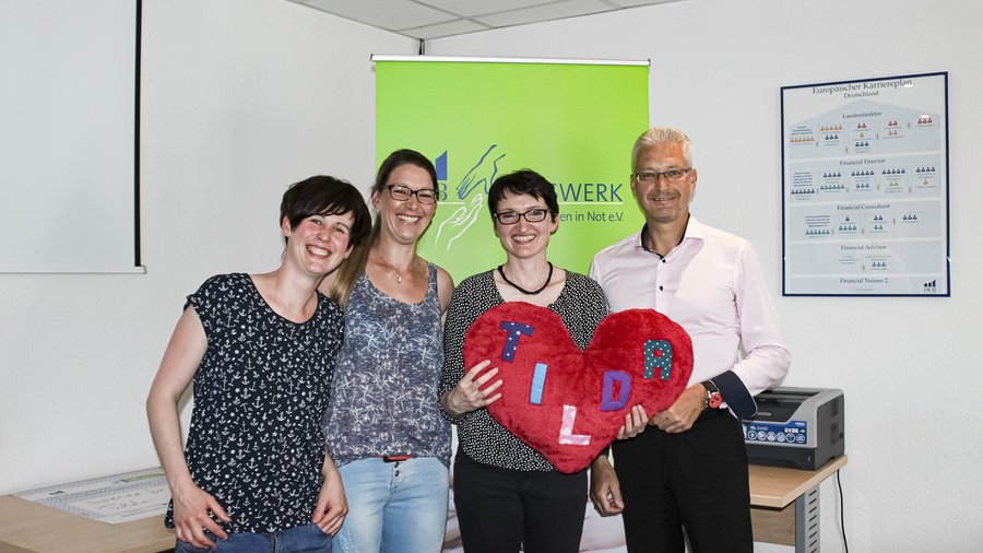 3 Frauen und ein Mann halten gemeinsam ein Herz hoch. Sie stehen zusammen im Büro und freuen sich