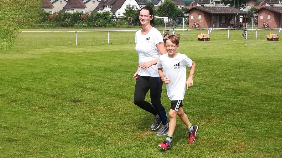 Frau und Kind laufen auf Wiese und blicken fröhlich in die Kamera bei Sommerwetter