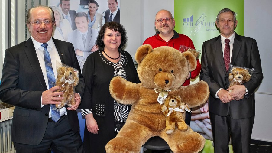 4 festlich gekleidete Menschen halten 3 Teddybären hoch. Ein Teddybär ist riesig und wird in der Mitte hochgehalten